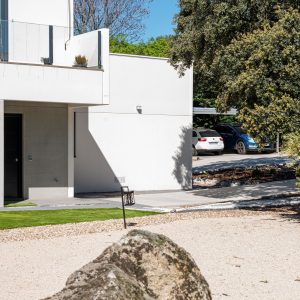Fotografía de la fachada lateral y acceso de la vivienda unifamiliar terminada en Galapagar, mostrando la materialización de los volúmenes de hormigón claro, la barandilla de vidrio del balcón y el jardín xerófilo de grava con una gran roca natural en primer plano, diseñada por Espacio Acorde.