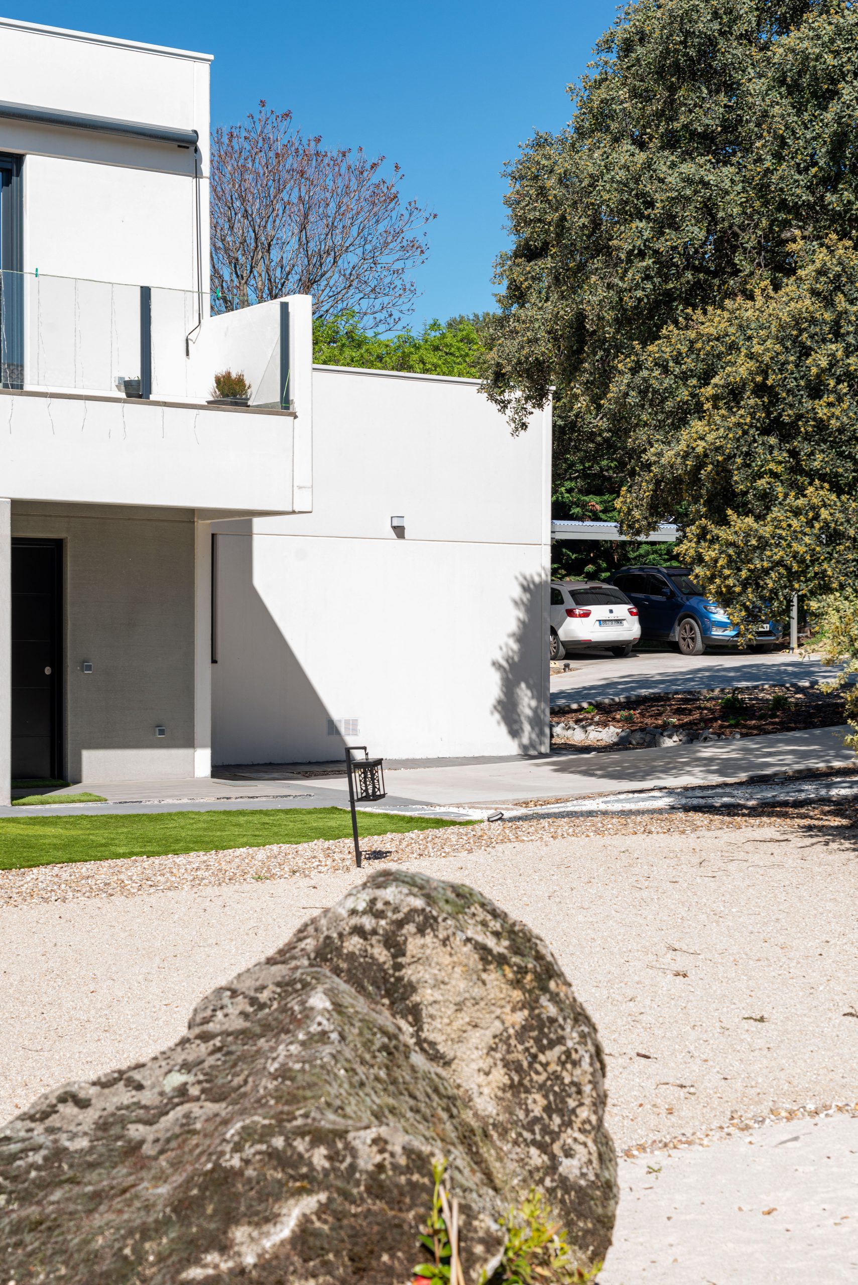 Fotografía de la fachada lateral y acceso de la vivienda unifamiliar terminada en Galapagar, mostrando la materialización de los volúmenes de hormigón claro, la barandilla de vidrio del balcón y el jardín xerófilo de grava con una gran roca natural en primer plano, diseñada por Espacio Acorde.