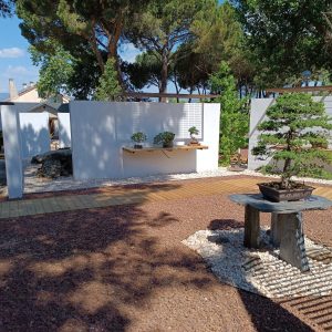 Patio de exposición exterior terminado en el Museo Municipal del Bonsái en Collado Villalba, mostrando los expositores de pizarra y madera, los lienzos de muros blancos, la grava volcánica y el pavimentado de madera