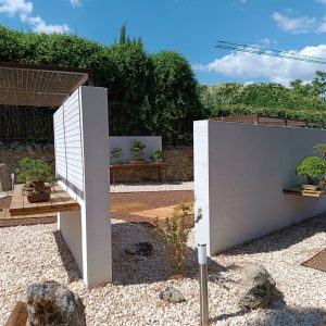 Museo Municipal del Bonsái en Collado Villalba mostrando la zonificación de patios mediante muros exentos blancos, pasarelas de madera natural, grava decorativa y balizas de iluminación.