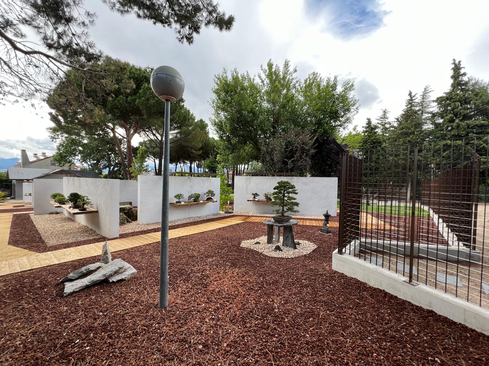 Vista panorámica del jardín exterior del Museo Municipal del Bonsái en Collado Villalba, mostrando la interacción entre las pasarelas de madera, el pavimento de corteza, los muros de exposición blancos y el vallado perimetral
