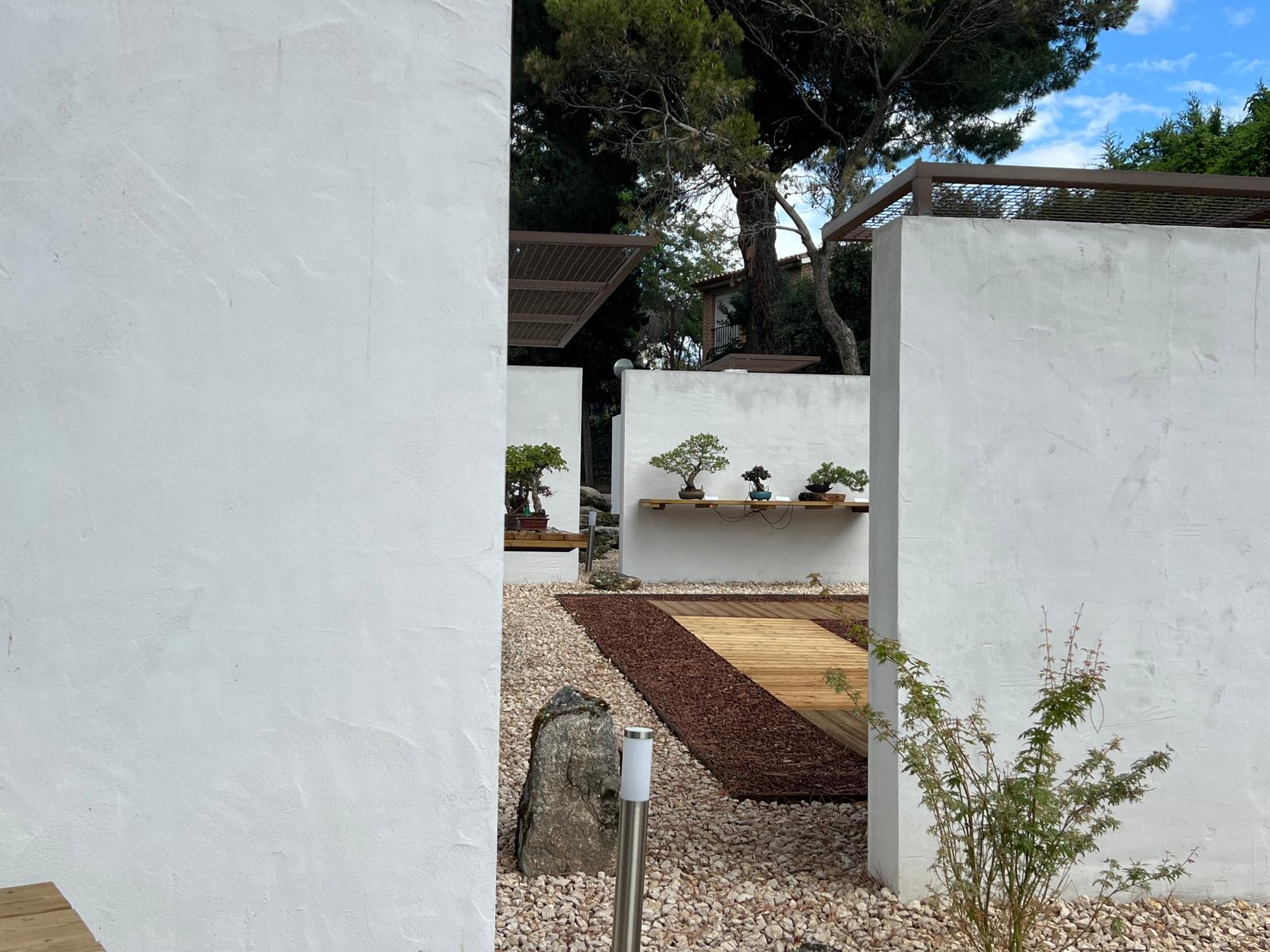 Fotografía de los patios exteriores del Museo del Bonsái en Collado Villalba, mostrando la transición entre muros blancos enfoscados, pasarelas de madera, grava decorativa y peanas de exposición