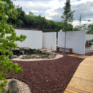 Panorámica del jardín exterior del Museo Municipal del Bonsái en Collado Villalba, mostrando la integración de pasarelas de madera tratada, pavimento de grava volcánica, un estanque con cascada de piedra natural y muros exentos blancos de exposición, diseñado por Espacio Acorde.