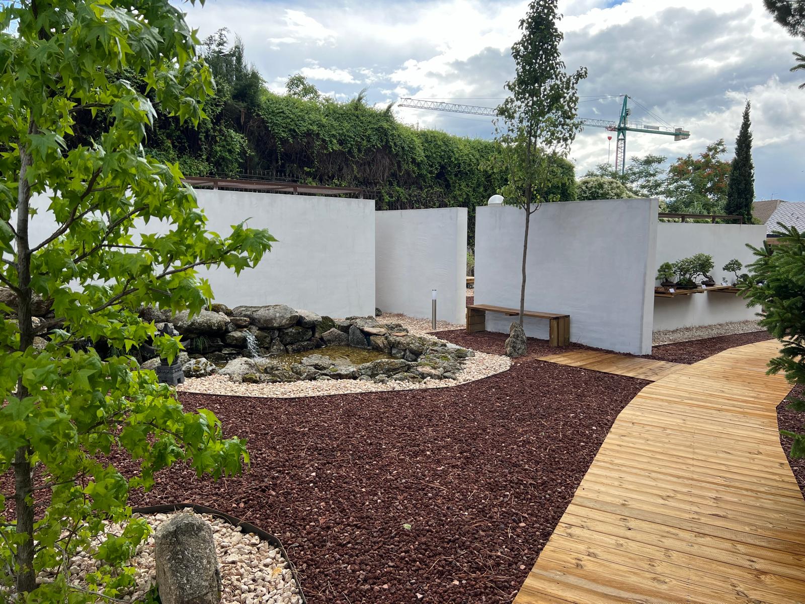 Panorámica del jardín exterior del Museo Municipal del Bonsái en Collado Villalba, mostrando la integración de pasarelas de madera tratada, pavimento de grava volcánica, un estanque con cascada de piedra natural y muros exentos blancos de exposición, diseñado por Espacio Acorde.