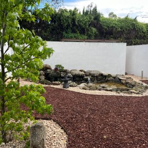 estanque con cascada de piedra natural, linterna de jardín zen de estilo japonés y un árbol joven integrados sobre grava volcánica y blanca frente a muros limpios en el Museo del Bonsái de Collado Villalba, diseñado por Espacio Acorde.