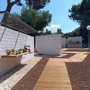 Pasarelas de madera y zonas de exposición del Museo Municipal del Bonsái en Collado Villalba, mostrando la integración de muros blancos, pérgolas de sombreado y pavimentos de grava volcánica, diseñado por Espacio Acorde.