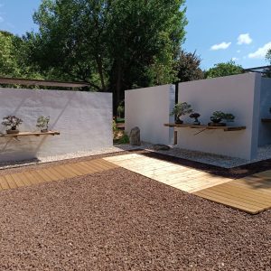 Patios de exposición al aire libre en el Museo Municipal del Bonsái en Collado Villalba, mostrando la convergencia de las pasarelas de madera, el pavimento de grava y los estantes de madera sobre muros blancos.