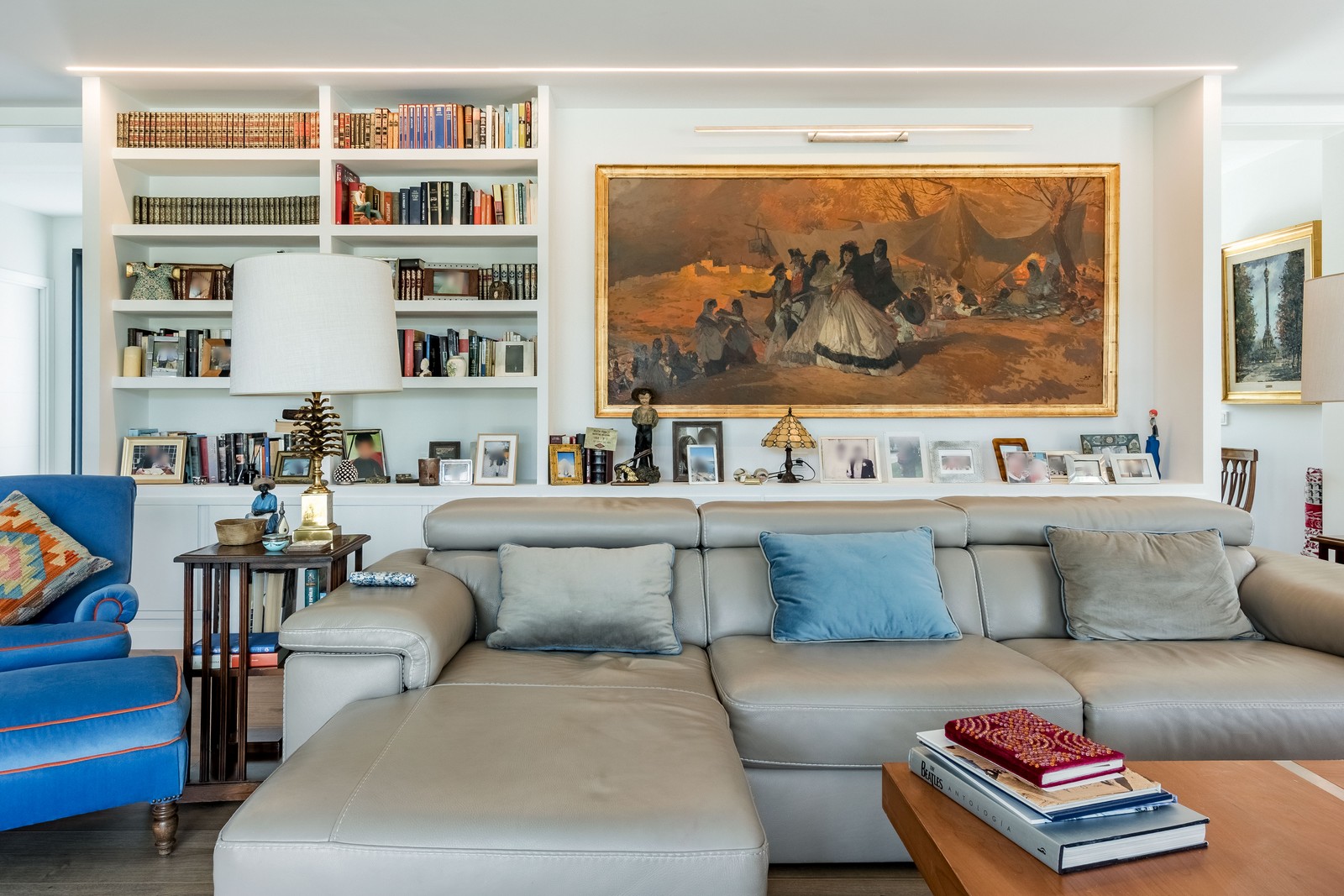 Zona de estar en salón de obra nueva en Madrid con biblioteca a medida integrada, iluminación LED perimetral, sofá gris, lienzo de gran formato y muebles clásicos.