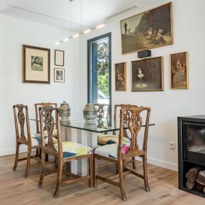 Zona de comedor con mesa de cristal, sillas de madera tallada, galería de cuadros mural y estufa de leña en vivienda unifamiliar de obra nueva en Madrid.