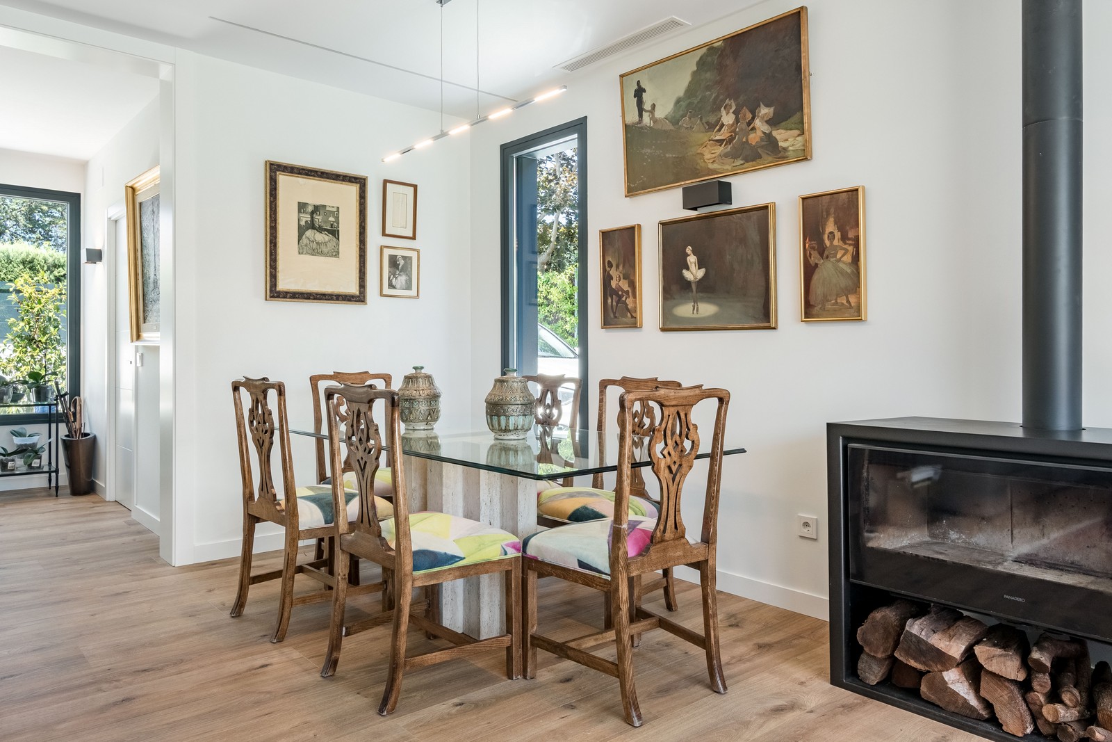Zona de comedor con mesa de cristal, sillas de madera tallada, galería de cuadros mural y estufa de leña en vivienda unifamiliar de obra nueva en Madrid.