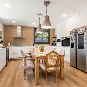 Vista panorámica de cocina y comedor de obra nueva en Madrid con revestimiento alistonado de madera, frente de trabajo en L, isla con mesa de madera maciza y pavimento continuo.
