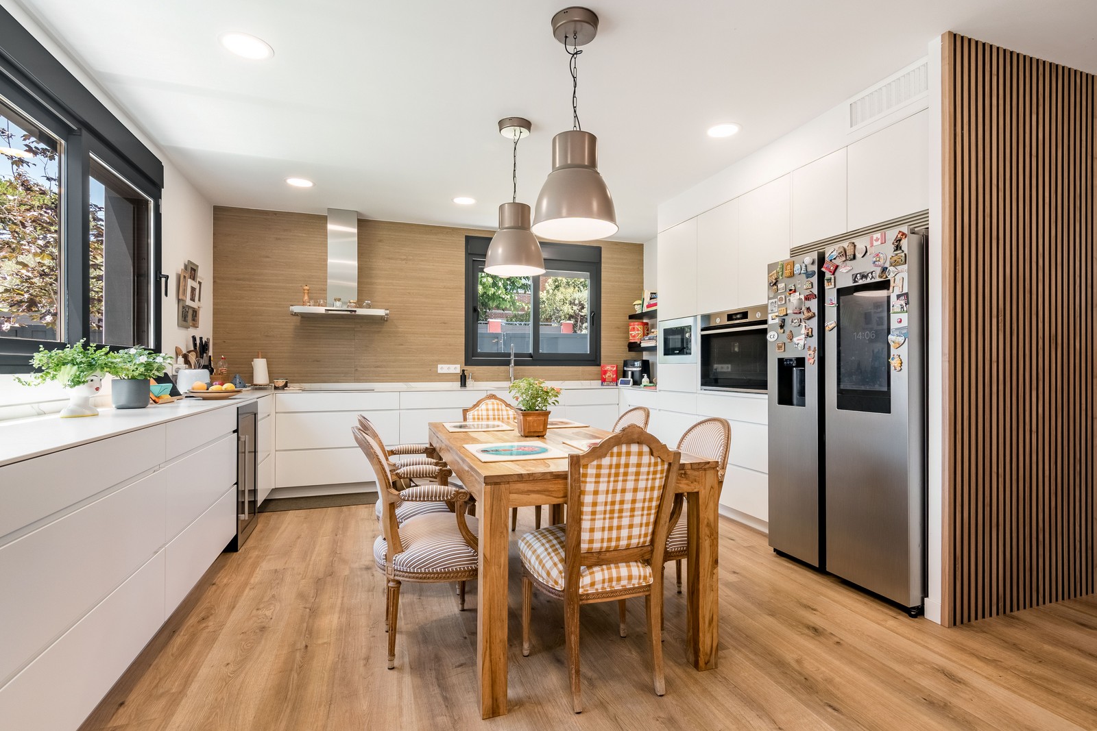 Vista panorámica de cocina y comedor de obra nueva en Madrid con revestimiento alistonado de madera, frente de trabajo en L, isla con mesa de madera maciza y pavimento continuo.
