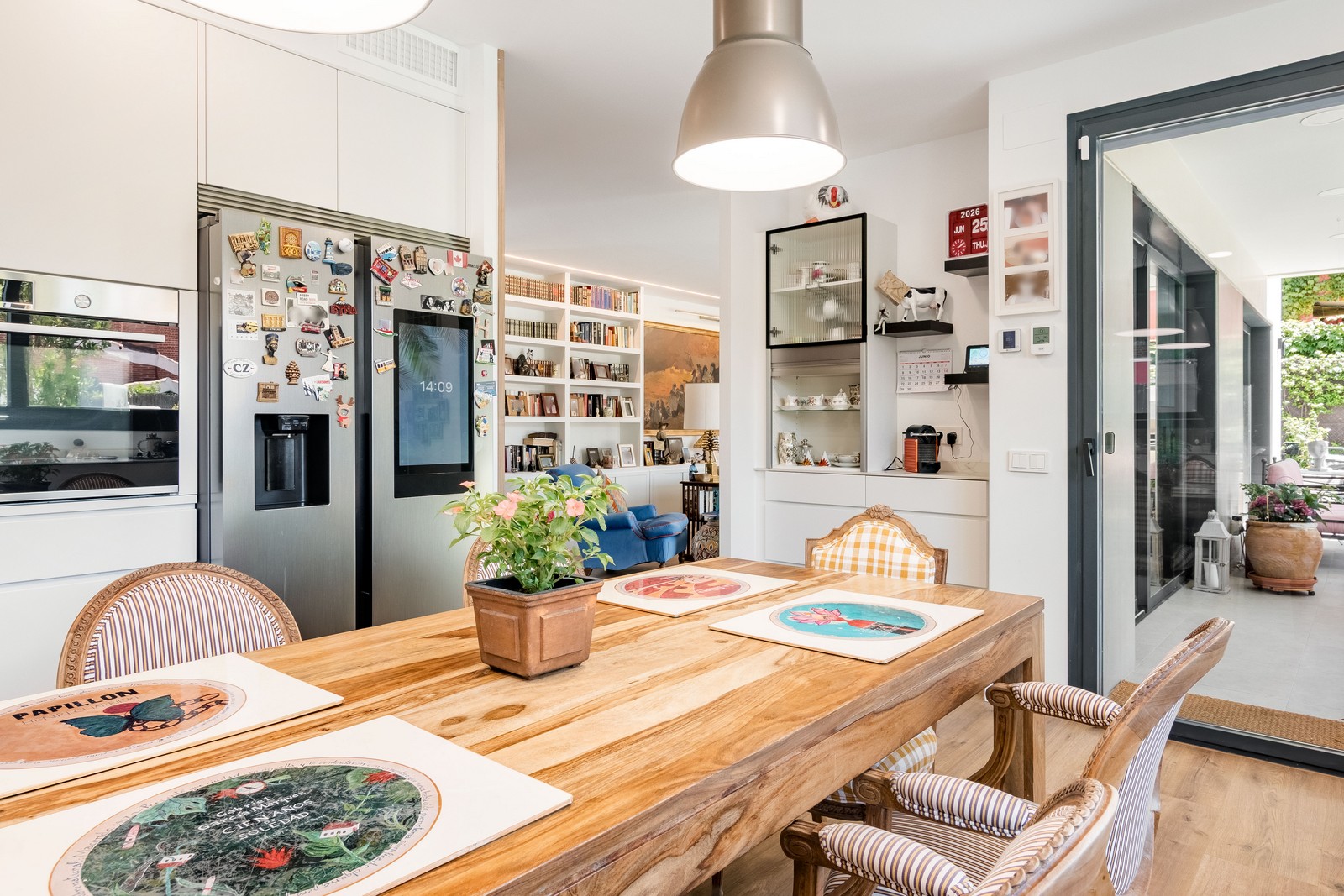 Vista de cocina y comedor de obra nueva en Madrid con mesa de madera maciza, electrodomésticos integrados, conexión directa al porche mediante gran ventanal y salón al fondo