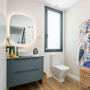 Aseo en vivienda unifamiliar de obra nueva en Madrid con mueble azul alistonado suspendido, espejo retroiluminado, ventana oscilobatiente y suelo cerámico de acabado madera.