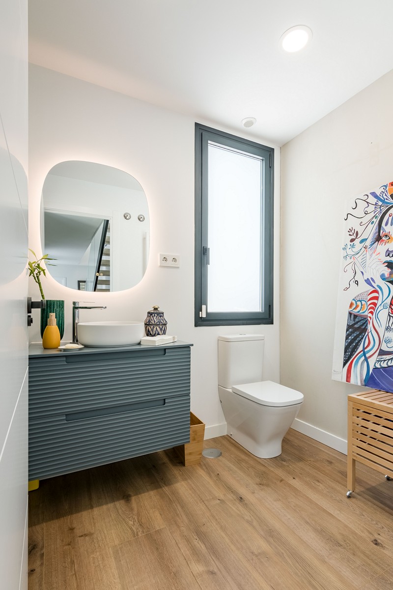 Aseo en vivienda unifamiliar de obra nueva en Madrid con mueble azul alistonado suspendido, espejo retroiluminado, ventana oscilobatiente y suelo cerámico de acabado madera.