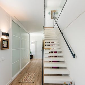 Recibidor de vivienda unifamiliar de obra nueva en Madrid con escalera volada blanca, armarios empotrados de vidrio translúcido y pavimento continuo.