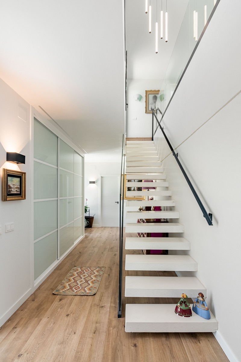 Recibidor de vivienda unifamiliar de obra nueva en Madrid con escalera volada blanca, armarios empotrados de vidrio translúcido y pavimento continuo.