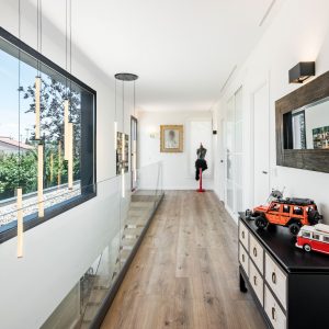 Distribuidor en doble altura de vivienda unifamiliar de obra nueva en Madrid con barandilla de vidrio, gran ventanal, lámparas suspendidas y pavimento continuo