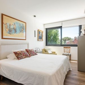 Vista amplia del dormitorio principal de obra nueva en Madrid, con gran ventanal, cabecero textil, zona de lectura y acceso con puerta de vidrio translúcido al baño.