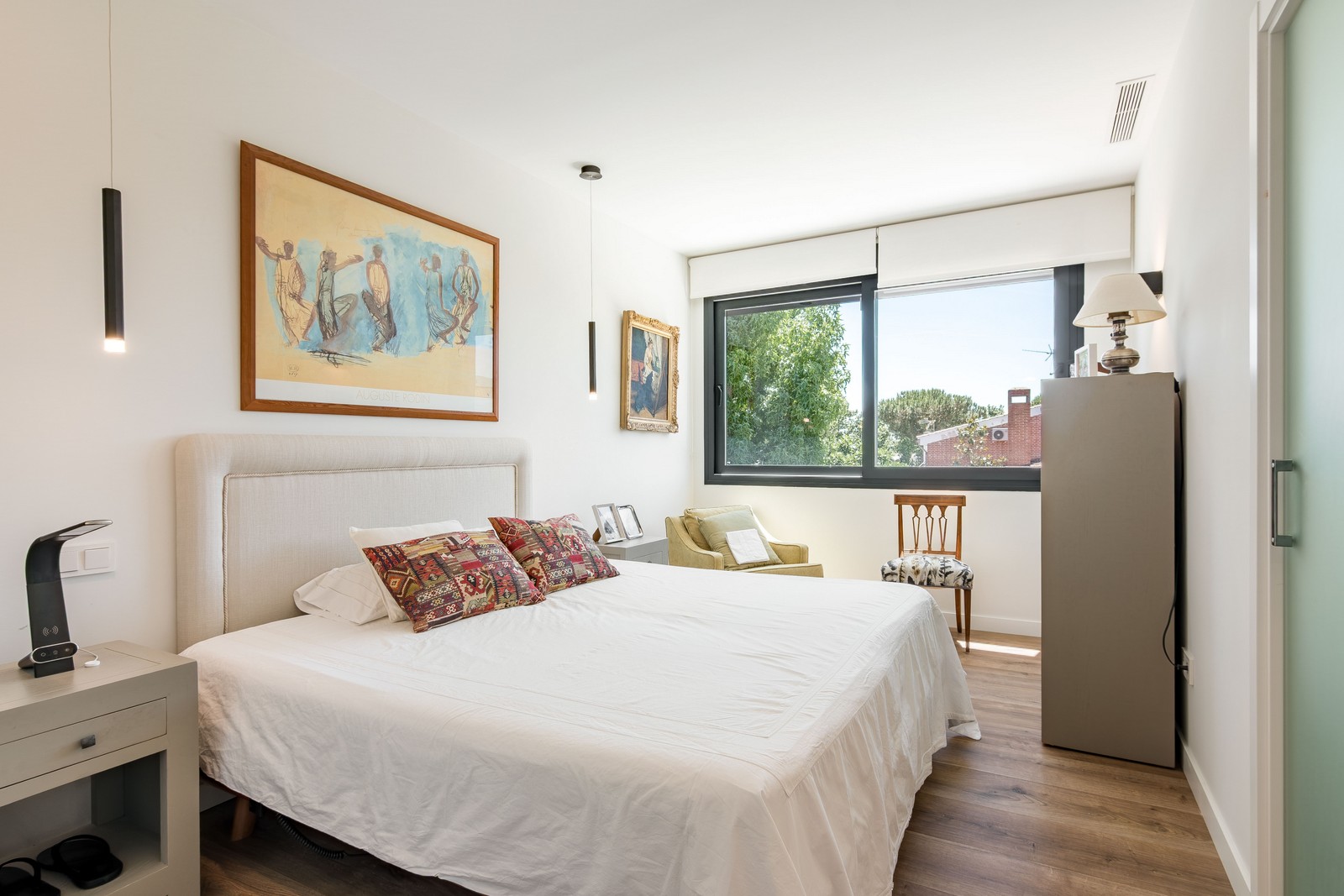 Vista amplia del dormitorio principal de obra nueva en Madrid, con gran ventanal, cabecero textil, zona de lectura y acceso con puerta de vidrio translúcido al baño.