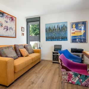 Diseño de despacho y sala polivalente en vivienda unifamiliar de obra nueva en Madrid, con escritorio frente a ventana, sofá mostaza, suelo laminado y luz natural.
