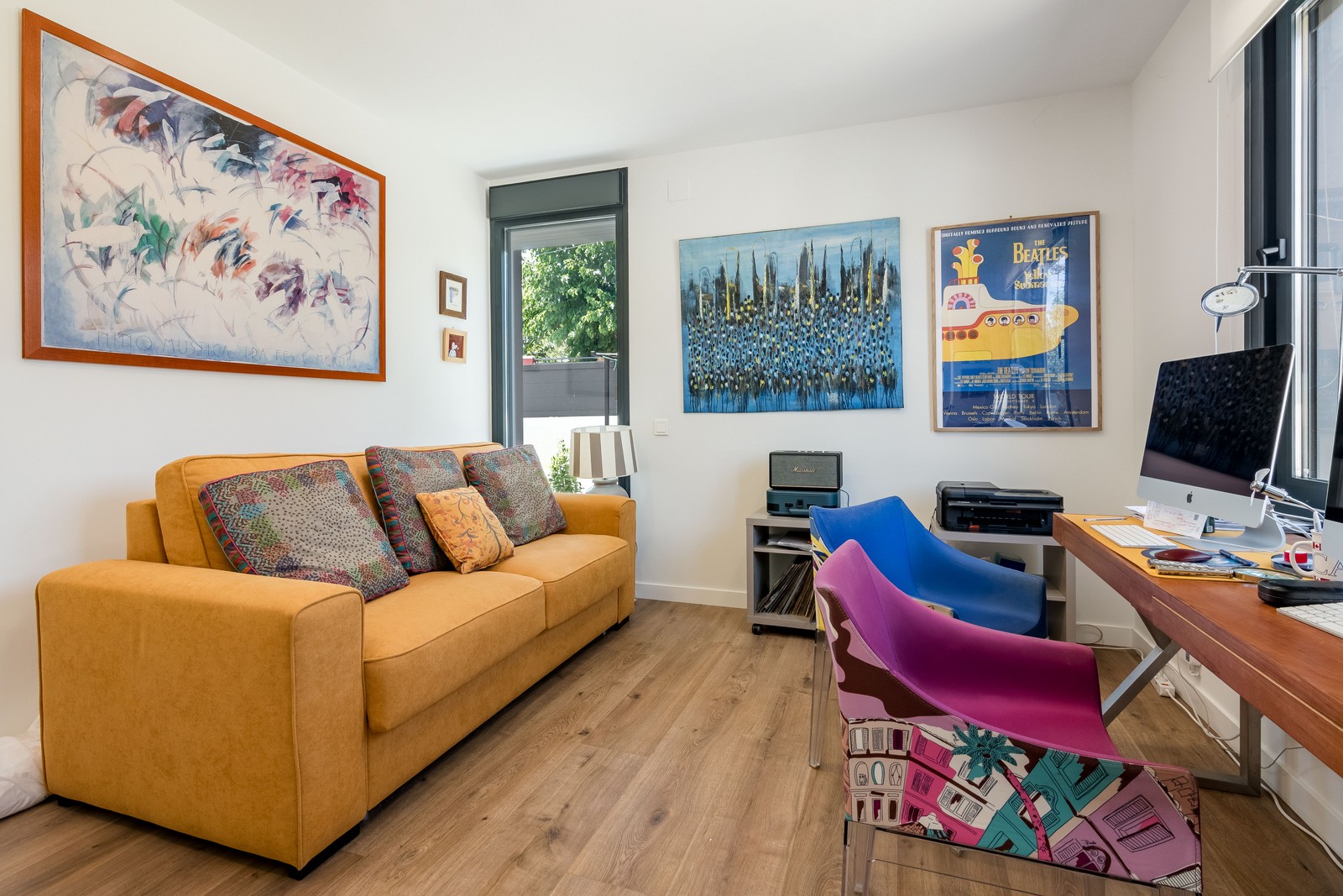 Diseño de despacho y sala polivalente en vivienda unifamiliar de obra nueva en Madrid, con escritorio frente a ventana, sofá mostaza, suelo laminado y luz natural.