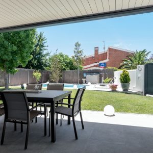 Vista desde el porche cubierto con comedor exterior hacia el jardín con césped y piscina privada en vivienda unifamiliar de obra nueva en Madrid.