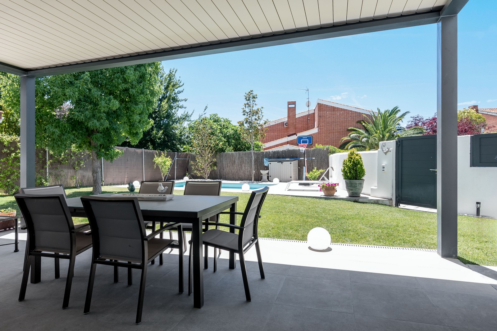 Vista desde el porche cubierto con comedor exterior hacia el jardín con césped y piscina privada en vivienda unifamiliar de obra nueva en Madrid.