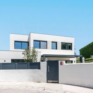 Vista frontal del acceso al jardín y porche de una vivienda unifamiliar de obra nueva en Madrid en blanco tiza y carpinterías gris grafito