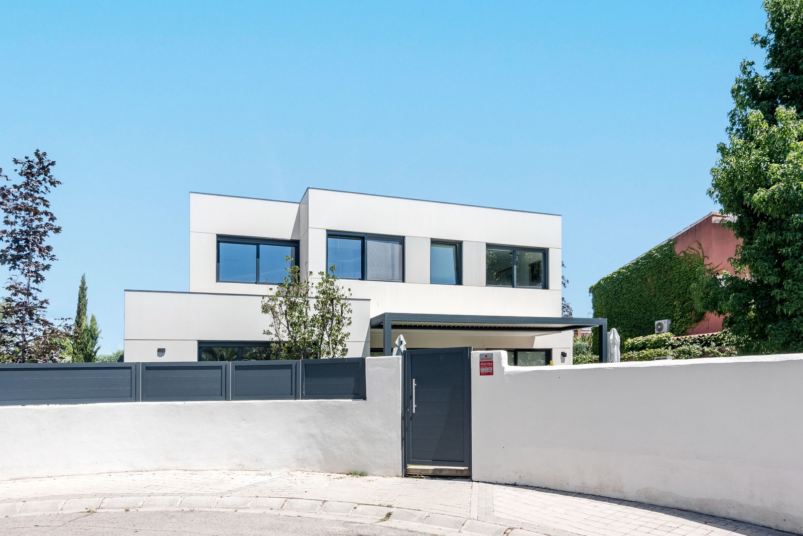 Vista frontal del acceso al jardín y porche de una vivienda unifamiliar de obra nueva en Madrid en blanco tiza y carpinterías gris grafito