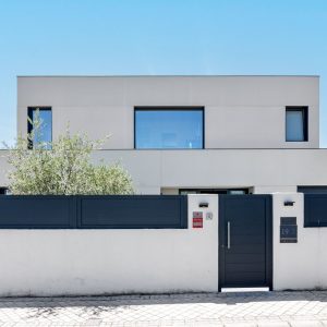 Fachada de estilo contemporáneo de obra nueva en Madrid con acabado SATE blanco, grandes ventanales de aluminio gris, muro perimetral con puerta de acceso y vegetación integrada.