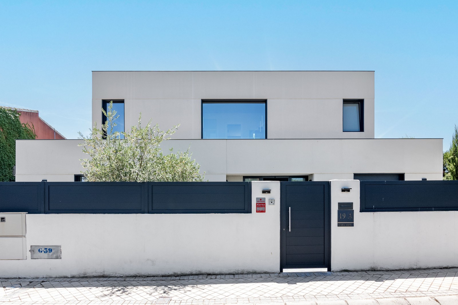 Fachada de estilo contemporáneo de obra nueva en Madrid con acabado SATE blanco, grandes ventanales de aluminio gris, muro perimetral con puerta de acceso y vegetación integrada.