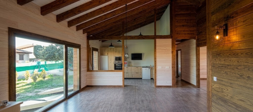 Interior luminoso de casa de madera en Colmenarejo con salón comedor de techos altos con vigas de madera vista y cocina semiabierta integrada.