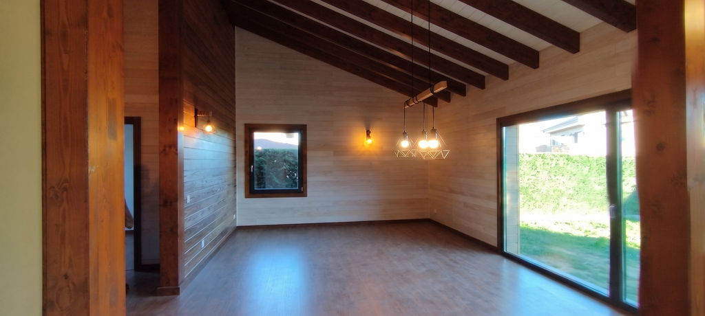Interior de salón diáfano con techo inclinado de vigas de madera vista, paredes revestidas de madera natural, iluminación cálida y grandes ventanales con vistas al jardín en Colmenarejo.