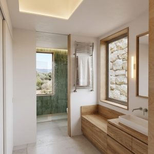 Vista de acceso a baño suite en ampliación en Torrelodones, con foso de iluminación cenital en techo, frentes de armario y mueble bajo en madera clara, ventanal hacia muro de piedra exterior y ducha con azulejo verde al fondo, por Espacio Acorde.