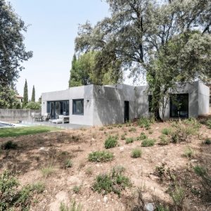 Vista general de la fachada de hormigón gris de una vivienda unifamiliar prefabricada en Galapagar, con piscina integrada en el jardín y rodeada de encinas centenarias en la sierra de Madrid, diseñada por Espacio Acorde.