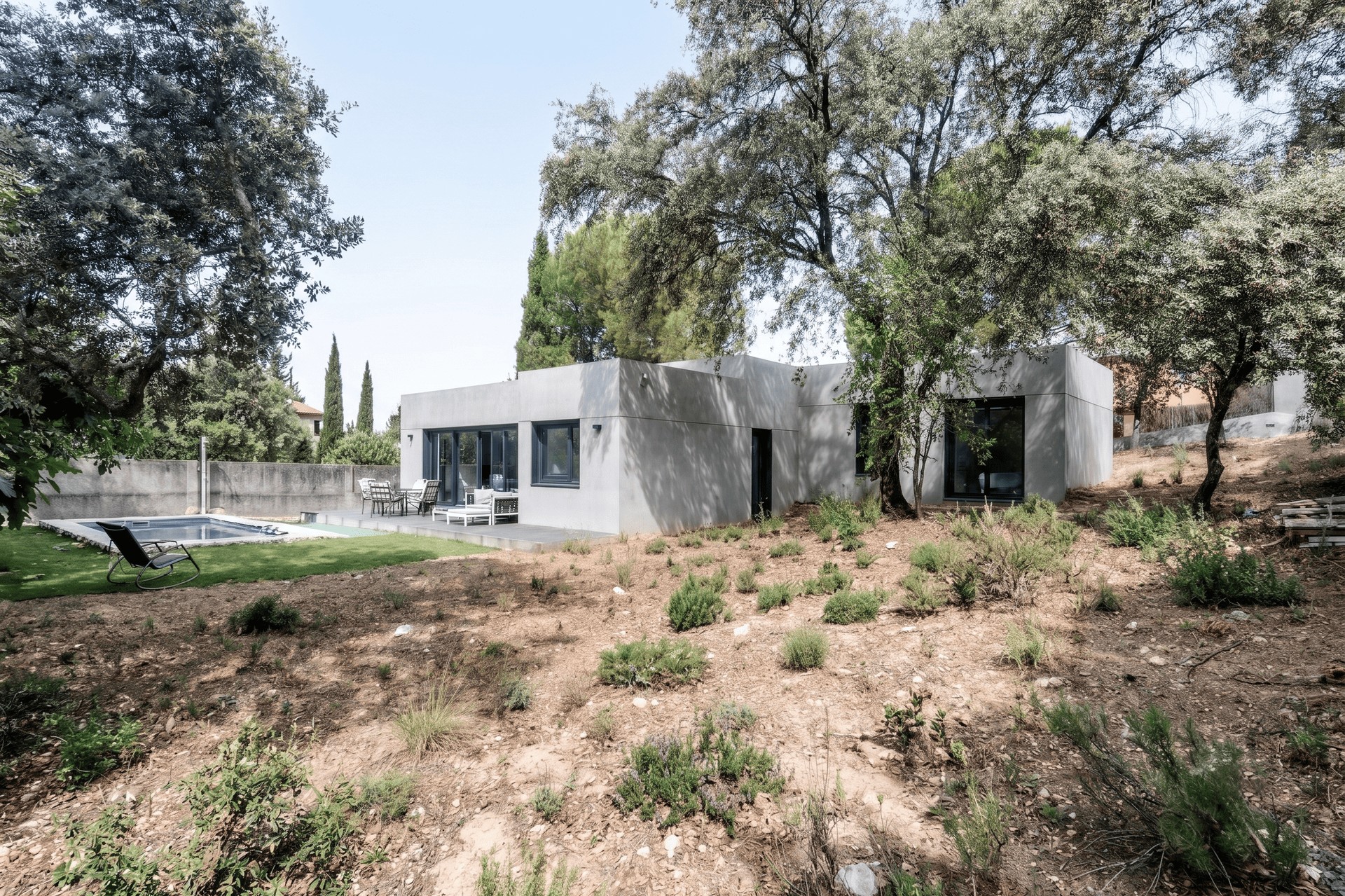 Vista general de la fachada de hormigón gris de una vivienda unifamiliar prefabricada en Galapagar, con piscina integrada en el jardín y rodeada de encinas centenarias en la sierra de Madrid, diseñada por Espacio Acorde.