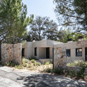 Vista desde la calle de una vivienda unifamiliar prefabricada de hormigón en la sierra de Madrid, con muros de cerramiento de piedra natural y pinos preservados, diseñada por Espacio Acorde