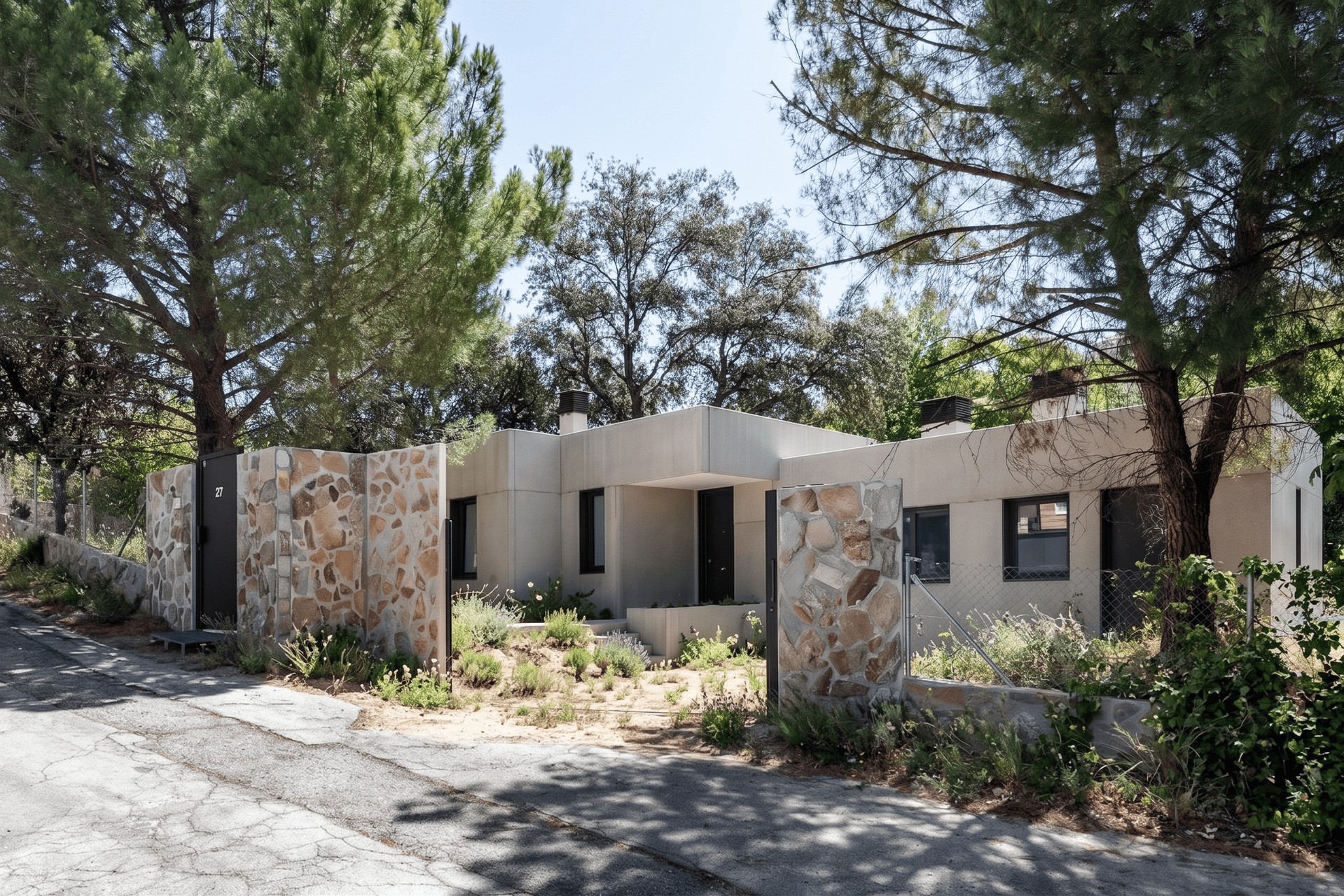 Vista desde la calle de una vivienda unifamiliar prefabricada de hormigón en la sierra de Madrid, con muros de cerramiento de piedra natural y pinos preservados, diseñada por Espacio Acorde
