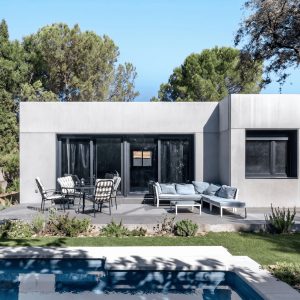 Fotografía de la fachada posterior de una vivienda unifamiliar prefabricada de hormigón en Madrid, mostrando una terraza pavimentada con zona de estar, comedor exterior y piscina en primer plano, diseñada por Espacio Acorde