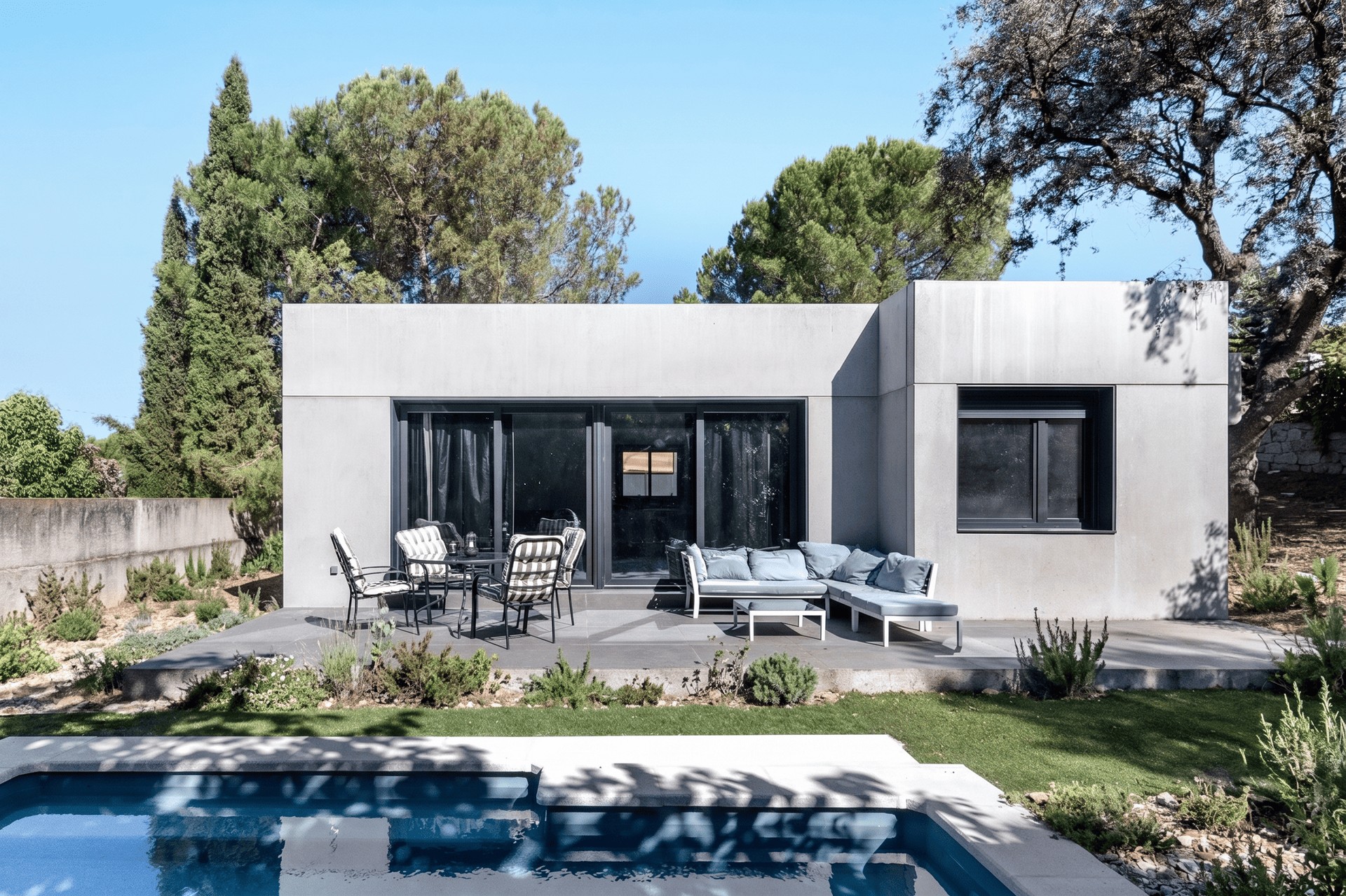 Fotografía de la fachada posterior de una vivienda unifamiliar prefabricada de hormigón en Madrid, mostrando una terraza pavimentada con zona de estar, comedor exterior y piscina en primer plano, diseñada por Espacio Acorde