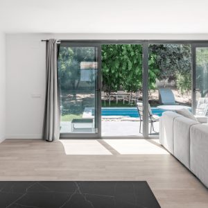 Vista interior de un salón moderno y luminoso con gran ventanal corredero que conecta con la piscina y el porche exterior, con suelo de madera clara y sofá gris, diseñado por Espacio Acorde