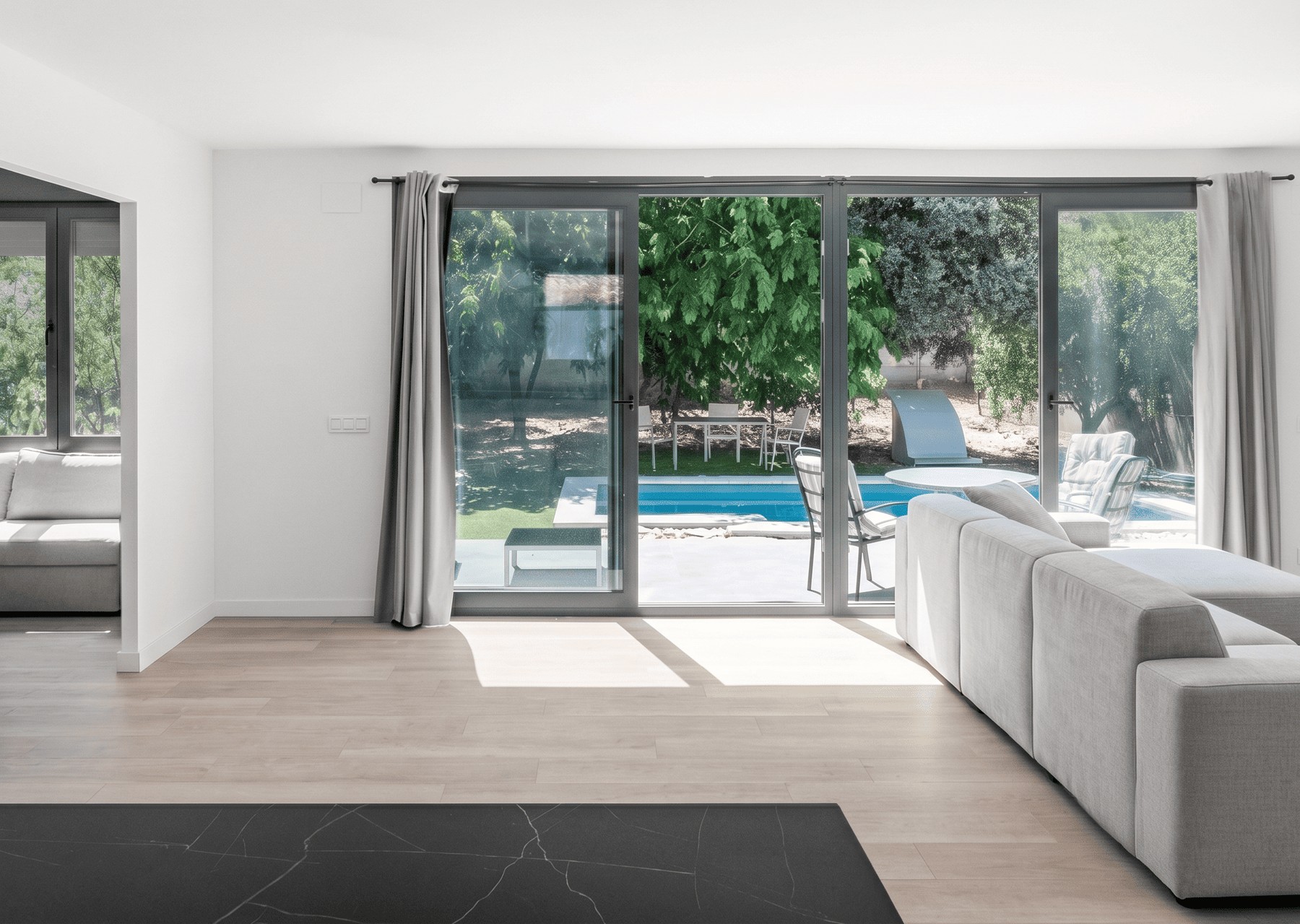 Vista interior de un salón moderno y luminoso con gran ventanal corredero que conecta con la piscina y el porche exterior, con suelo de madera clara y sofá gris, diseñado por Espacio Acorde