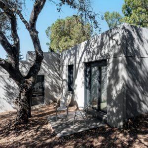 Fachada posterior de una vivienda unifamiliar prefabricada de hormigón en Madrid, mostrando un rincón de descanso con dos sillas metálicas bajo la sombra de una encina centenaria que proyecta siluetas sobre el muro gris, diseñada por Espacio Acorde.