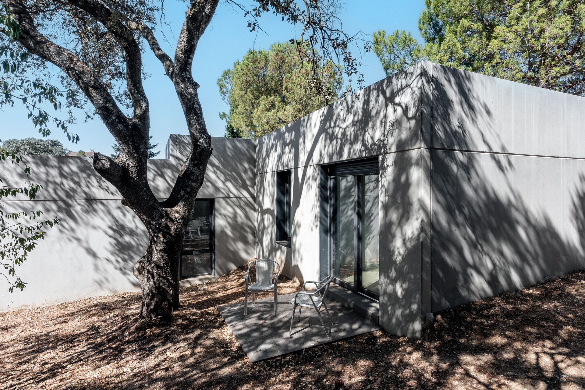 Fachada posterior de una vivienda unifamiliar prefabricada de hormigón en Madrid, mostrando un rincón de descanso con dos sillas metálicas bajo la sombra de una encina centenaria que proyecta siluetas sobre el muro gris, diseñada por Espacio Acorde.