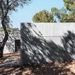 Fotografía exterior de una casa unifamiliar prefabricada de hormigón gris en Madrid, mostrando el juego de luces y sombras proyectadas por los árboles sobre la fachada y la conexión con la terraza exterior, diseñada por Espacio Acorde.