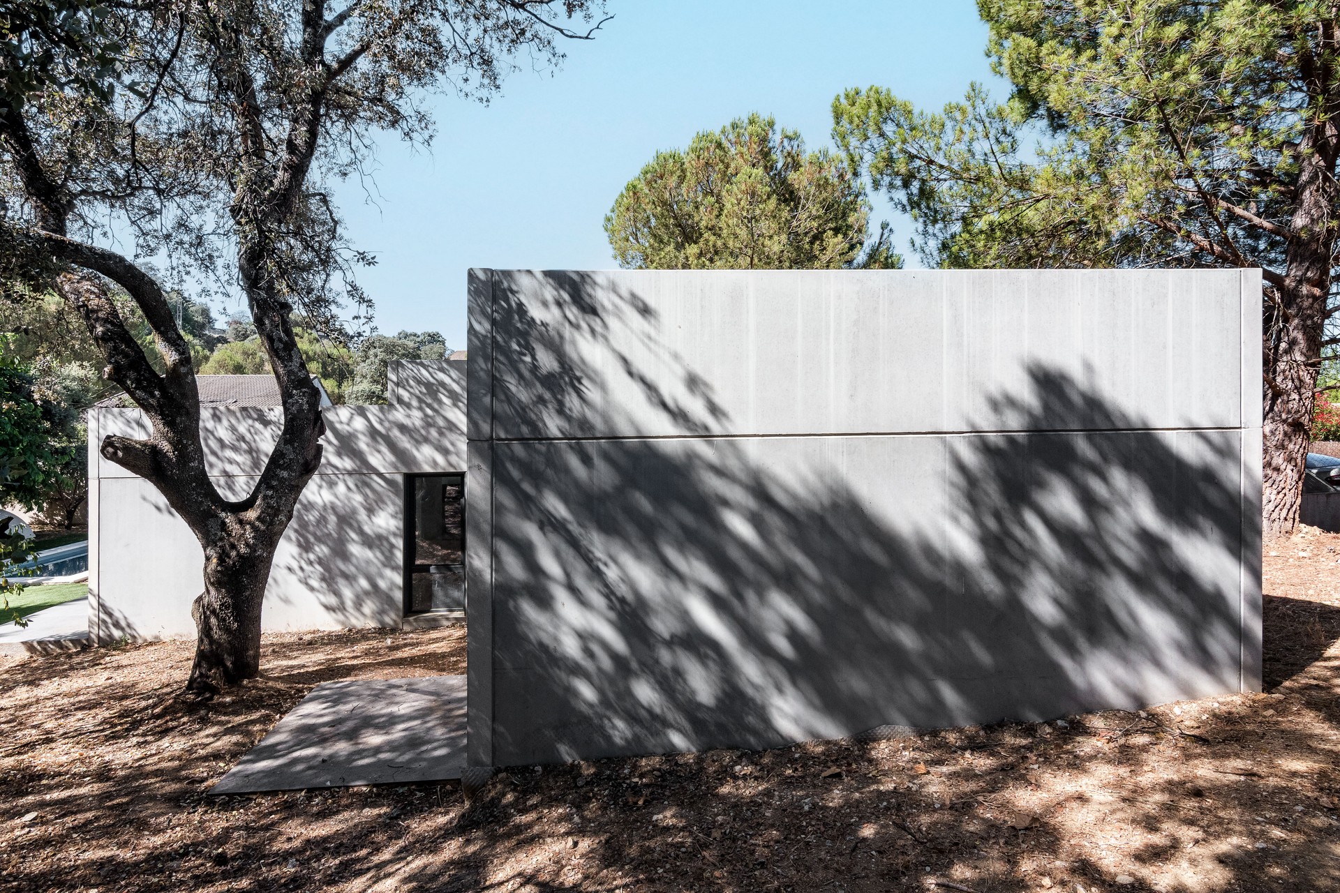 Fotografía exterior de una casa unifamiliar prefabricada de hormigón gris en Madrid, mostrando el juego de luces y sombras proyectadas por los árboles sobre la fachada y la conexión con la terraza exterior, diseñada por Espacio Acorde.