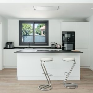 Vista interior de una cocina moderna con isla blanca y encimera negra en una vivienda unifamiliar en Galapagar, con ventana de aluminio oscuro y suelo de tarima de madera clara, diseñada por Espacio Acorde