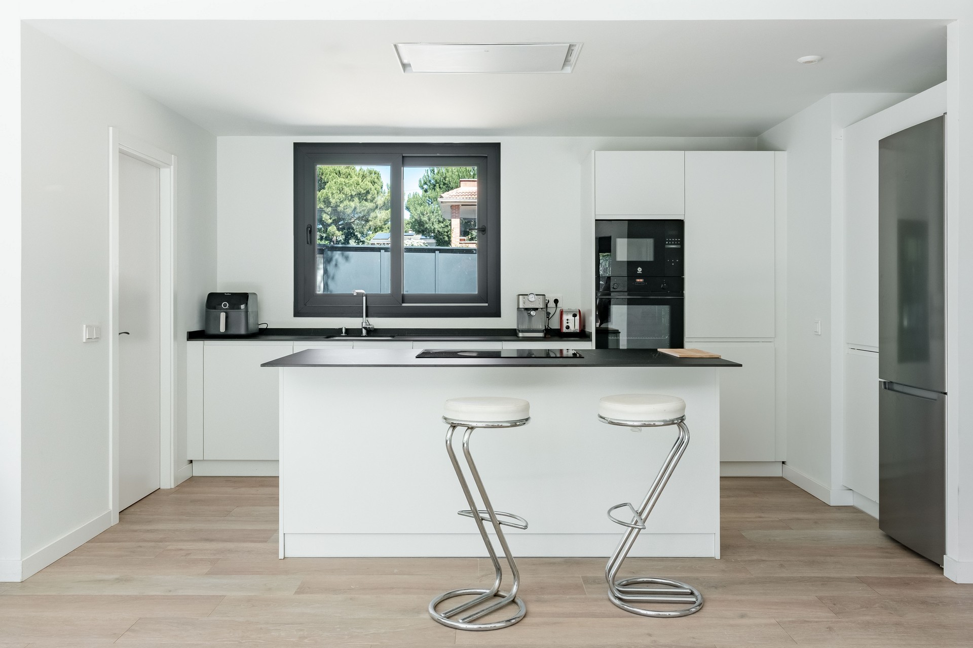 Vista interior de una cocina moderna con isla blanca y encimera negra en una vivienda unifamiliar en Galapagar, con ventana de aluminio oscuro y suelo de tarima de madera clara, diseñada por Espacio Acorde