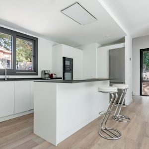 Perspectiva de cocina moderna blanca con isla en Galapagar, mostrando el flujo visual directo hacia el jardín natural a través de una puerta acristalada oscilobatiente, diseñada por Espacio Acorde.